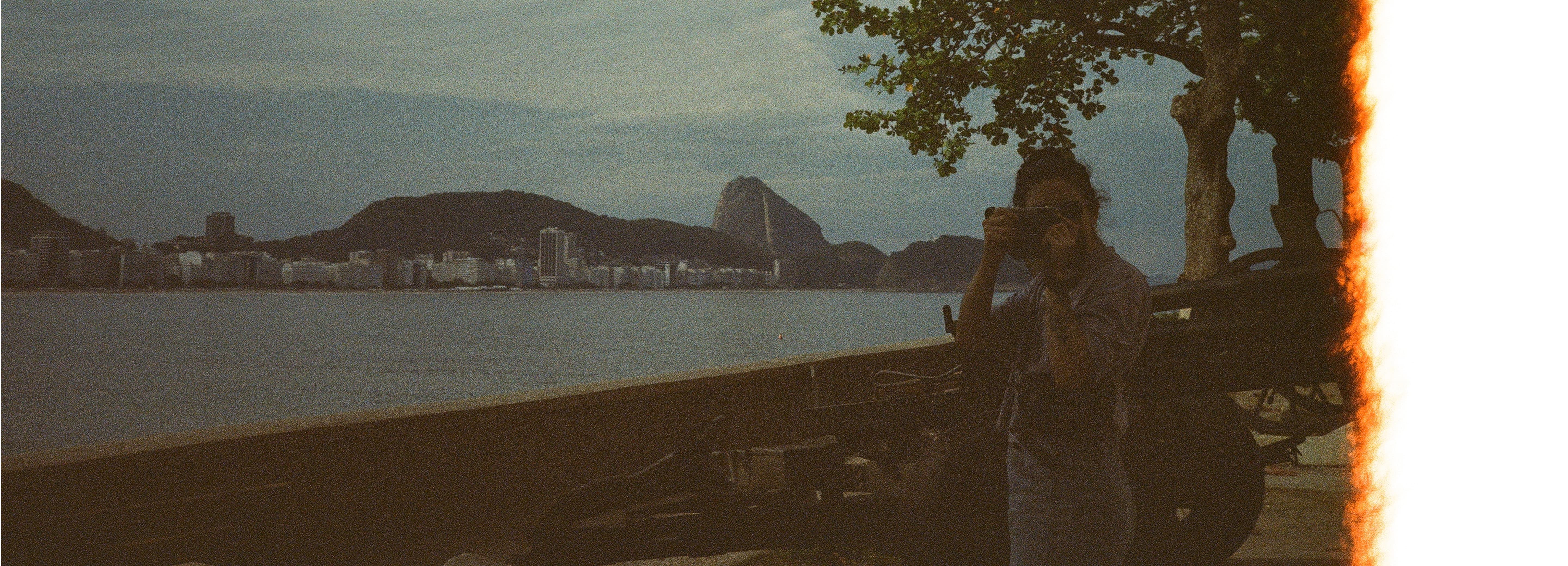 Grainy image of Luiza in a background landscape with a sea viewport and mountains. She is pointing a camera back to the photographer.